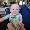baby, child, smiling, happy, onesie, sitting, person, indoor, furniture, cardboard_box, home, living_room, fan, clock, painting, plant, table, chair, door, window