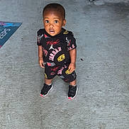 Kamari is registered to the contest to win money with this photo: toddler, child, concrete, floor, stairs, clothing, sneakers, curious, standing, looking_up, outdoor, doormat, black, red, yellow, patterned, young_child, person, casual, daylight