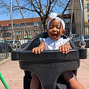 Ravyn joined the competition — help win amazing prizes! baby, child, daylight, feet, fence, happy, headband, metal, outdoor, park, person, playground, seat, smiling, sunny, swing, toddler, tree, urban, white_clothing