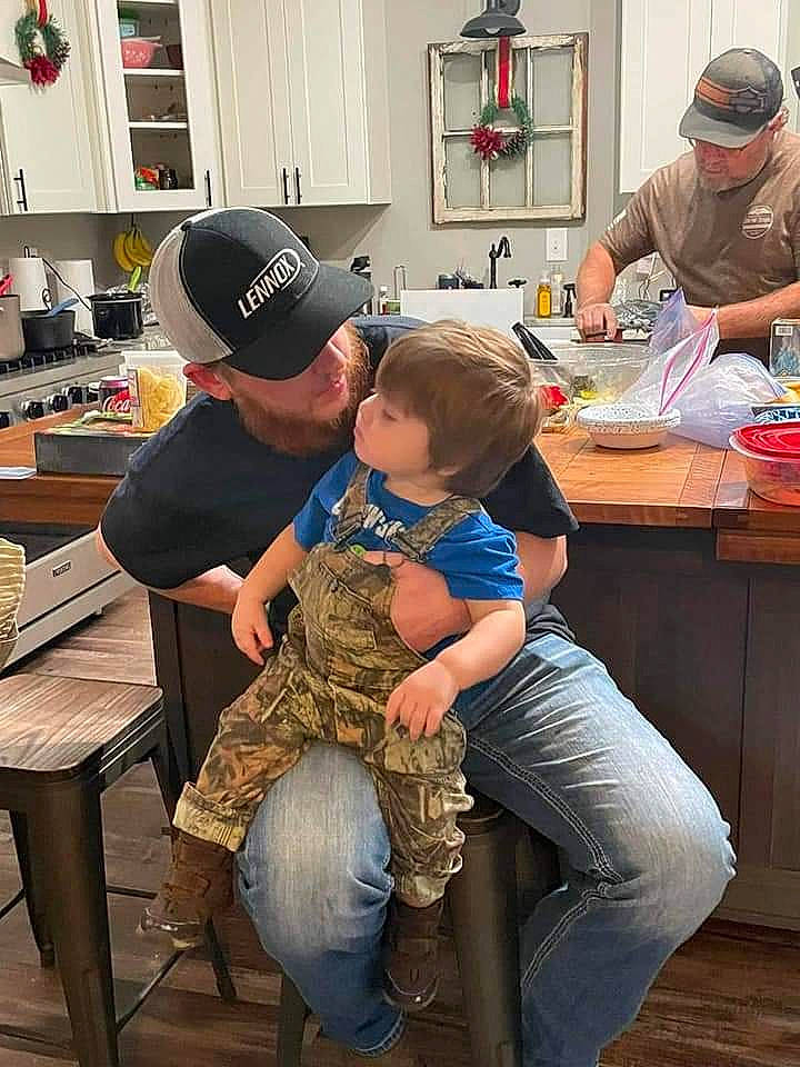 Dallas is registered to the contest to win money with this photo: arm, baseball_cap, cap, child, clothing, cooking, countertop, event, eyewear, flooring, fun, hat, headwear, jeans, kitchen, person, room, shelf, sitting, t_shirt