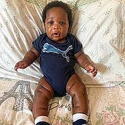 Zahari is registered to the contest to win money with this photo: baby, infant, child, onesie, puma_logo, socks, bedspread, floral_pattern, eiffel_tower, pillow, indoor, person, cute, sitting, curious, skin, face, head, legs, arm