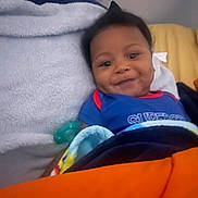 Braylin is registered to the contest to win money with this photo: baby, infant, child, smile, dimple, blanket, pacifier, bed, towel, onesie, blue_clothing, orange_blanket, pillow, face, hair, cozy, indoors, lying_down, closeup, happy