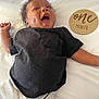 baby, infant, smiling, laughing, one_month, milestone_disc, wooden_sign, black_shirt, tshirt, blanket, lying_down, chubby_cheeks, hand, open_mouth, hair, skin, portrait, happy, bed, cute