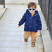 Marius a rejoint le concours — aidez-le/la à gagner de superbes lots ! child, toddler, sunglasses, navy_coat, jacket, brown_pants, boots, walking, outdoor, tiled_floor, railing, concrete_wall, portrait, cute, smile, winter_clothing, fashion, pavement, blue_sunglasses, urban