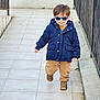 child, toddler, sunglasses, navy_coat, jacket, brown_pants, boots, walking, outdoor, tiled_floor, railing, concrete_wall, portrait, cute, smile, winter_clothing, fashion, pavement, blue_sunglasses, urban