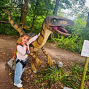 Hiba participe au concours pour gagner de l'argent avec cette photo : braided_hair, casual_clothing, child, daylight, dinosaur_statue, forest, girl, harness, jeans, leaves, nature, outdoor, park, pathway, plants, playground, sculpture, sneakers, toy, trees