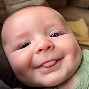 Finnley is registered to the contest to win money with this photo: baby, close_up, face, tongue, cheeks, blue_eyes, green_clothing, pacifier_clip, smiling, cute, infant, child, indoors, soft_texture, blanket, happy, person, portrait, young_child, skin