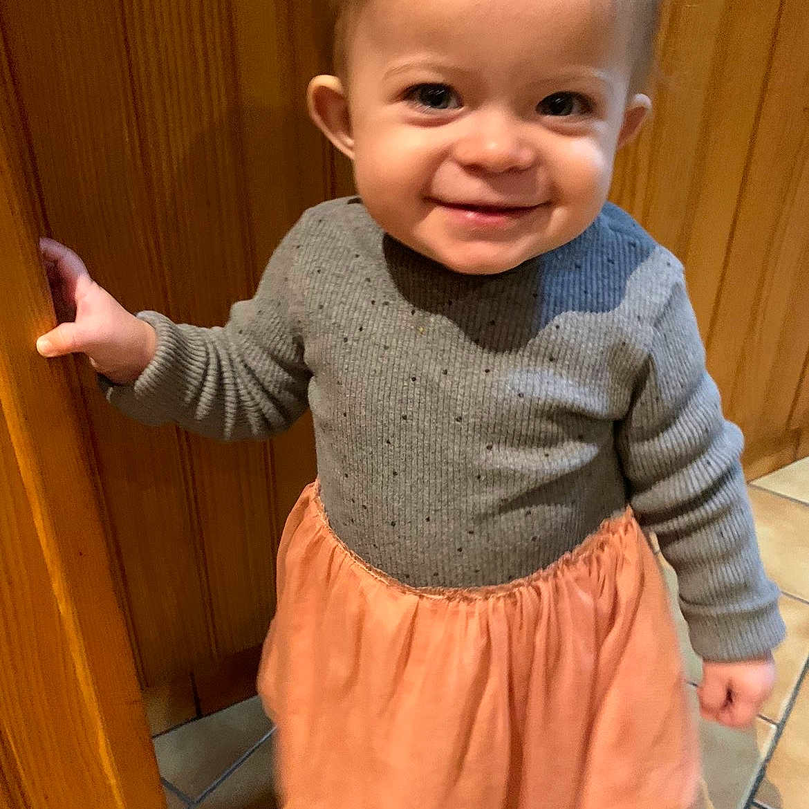 Zeze participe au concours pour gagner de l'argent avec cette photo : bright_eyes, casual, child, clothing, cute, floor_tiles, gray_sweater, hand_on_door, happy, indoor, peach_skirt, person, portrait, short_hair, smiling, standing, toddler, warm_lighting, wooden_door, young_child