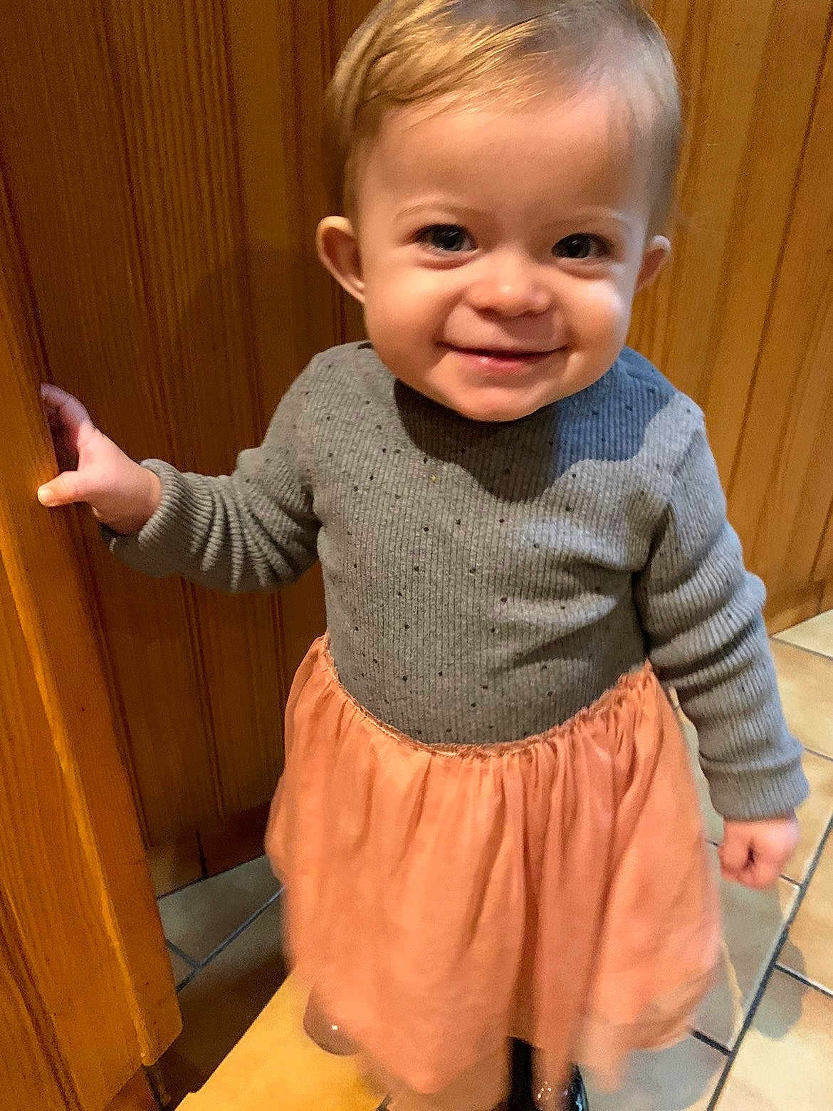 Zeze participe au concours pour gagner de l'argent avec cette photo : toddler, child, smiling, indoor, wooden_door, gray_sweater, peach_skirt, floor_tiles, happy, person, cute, young_child, portrait, standing, clothing, hand_on_door, bright_eyes, short_hair, warm_lighting, casual