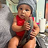 Kailo is registered to the contest to win money with this photo: baby, child, knitted_hat, horned_hat, red_clothing, drinking_horn, kitchen, person, holding, curious, cute, indoor, adult_hand, table, paper_towel, face, expression, sitting, skin, portrait