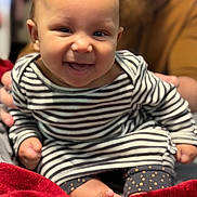 Kailo is registered to the contest to win money with this photo: baby, child, smiling, striped_clothing, sitting, red_blanket, indoor, person, happy, cute, infant, face, portrait, human, young_child, cozy, blurred_background, warm_colors, hands, feet