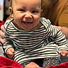 baby, child, smiling, striped_clothing, sitting, red_blanket, indoor, person, happy, cute, infant, face, portrait, human, young_child, cozy, blurred_background, warm_colors, hands, feet