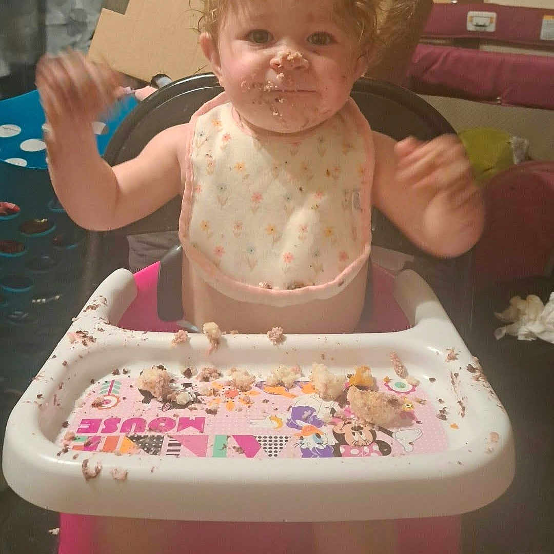 Kaylanna is registered to the contest to win money with this photo: baby, child, high_chair, eating, cake, crumbs, messy, smiling, waving, curly_hair, bib, infant, portrait, indoor, home, kitchen, tray, food, celebration, happy