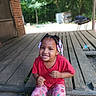 child, smiling, girl, braids, beads, red_shirt, pink_pants, patterned_clothing, wooden_porch, outdoor, daylight, greenery, brick_wall, crouching, happy, person, casual_clothing, nature, portrait, cute
