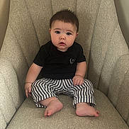 Dutton is registered to the contest to win money with this photo: baby, child, infant, armchair, striped_pants, black_shirt, barefoot, seated, indoors, cute, curious_expression, chubby_cheeks, furniture, home, portrait, person, young, small_hands, small_feet, casual_clothing