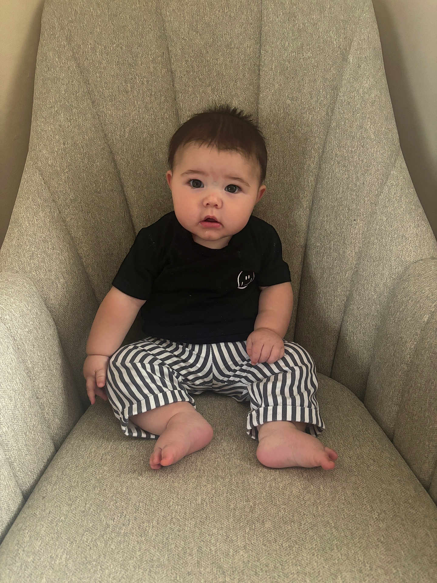 Dutton is registered to the contest to win money with this photo: baby, child, infant, armchair, striped_pants, black_shirt, barefoot, seated, indoors, cute, curious_expression, chubby_cheeks, furniture, home, portrait, person, young, small_hands, small_feet, casual_clothing