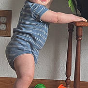 Marcus joined the competition — help win amazing prizes! baby, child, standing, onesie, striped_clothing, wooden_floor, wooden_table, toy, green_toy, orange_toy, knitted_blanket, indoor, smiling, happy, face, feet, hand, plant, wall_outlet, home