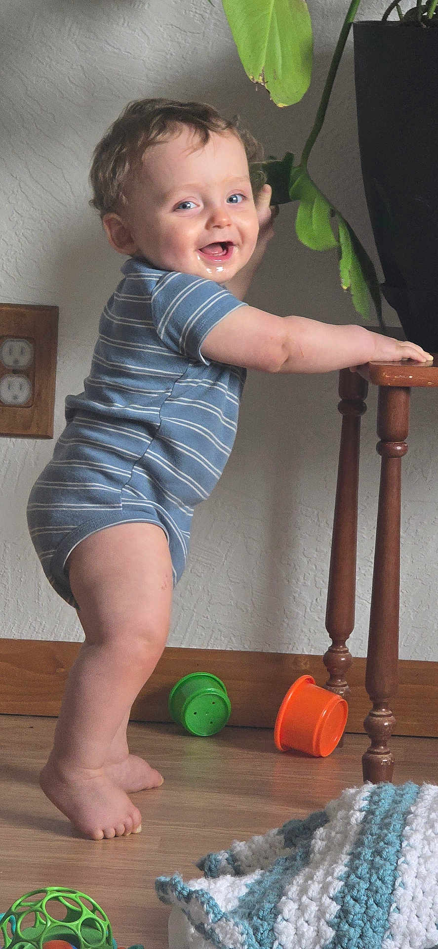 Marcus joined the competition — help win amazing prizes! baby, child, standing, onesie, striped_clothing, wooden_floor, wooden_table, toy, green_toy, orange_toy, knitted_blanket, indoor, smiling, happy, face, feet, hand, plant, wall_outlet, home