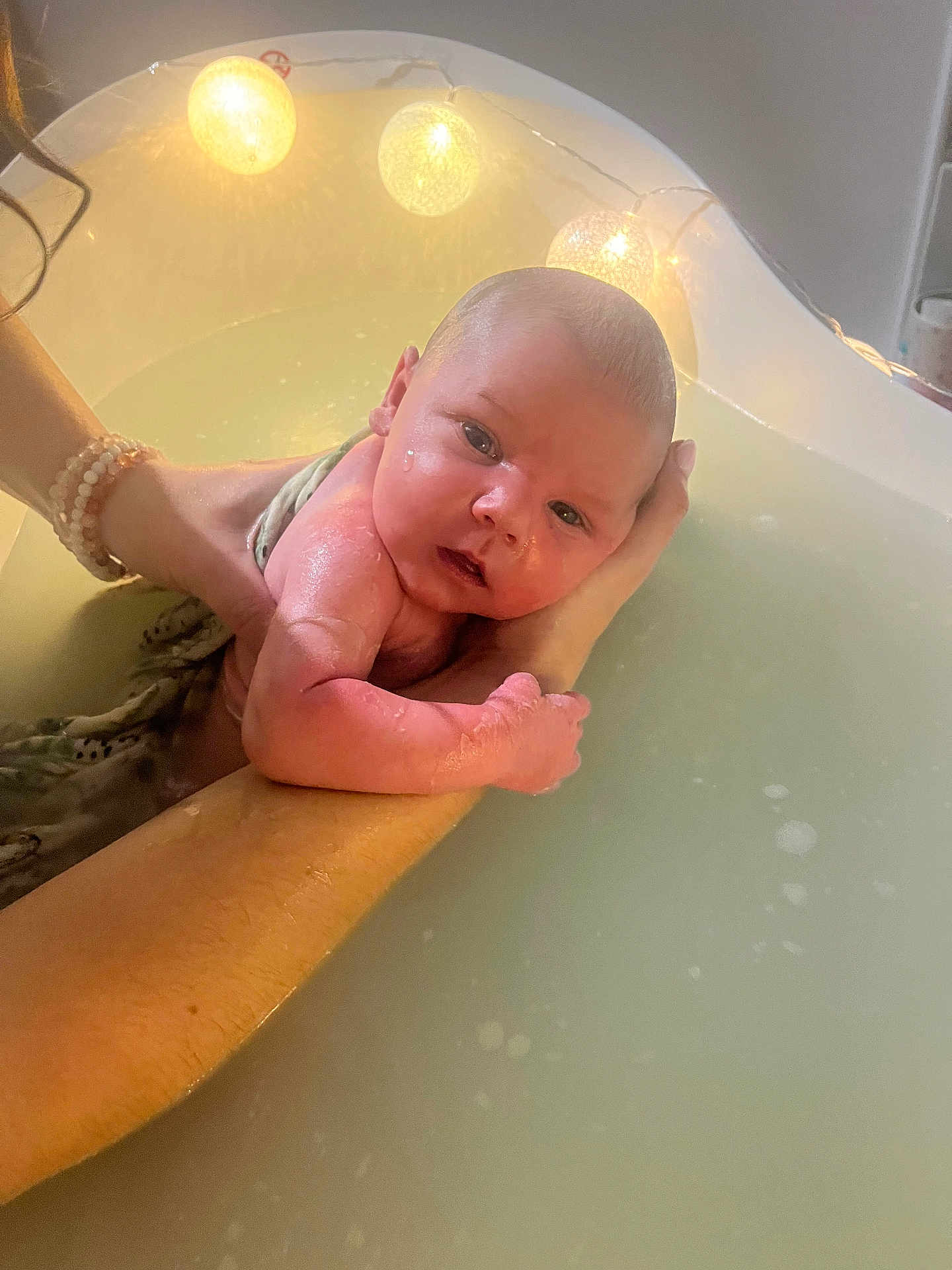 Ezio participe au concours pour gagner de l'argent avec cette photo : baby, infant, bath, water, hands, string_lights, tub, skin, face, person, newborn, arm, wet, soothing, comfort, indoor, lighting, closeup, parent, care