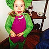 child, costume, green_costume, leaf_headpiece, cartoon, smiling, portrait, standing, indoors, wooden_floor, shelf, furniture, blue_chair, shoes, long_hair, colorful_clothing, playful, happy, full_body, family_photo