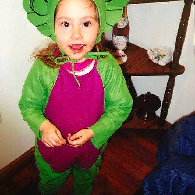 Katie joined the competition — help win amazing prizes! child, costume, green_costume, leaf_headpiece, cartoon, smiling, portrait, standing, indoors, wooden_floor, shelf, furniture, blue_chair, shoes, long_hair, colorful_clothing, playful, happy, full_body, family_photo