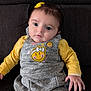 baby, child, infant, yellow_clothing, grey_dress, smiley_face, bow_hairclip, sitting, couch, indoor, portrait, cute, face, hand, long_sleeves, leggings, expression, young_child, kid, apparel