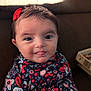 baby, child, face, tongue, smile, red_bow, floral_outfit, patterned_clothing, couch, indoor, portrait, cute, happy, infant, person, head, young_child, closeup, seated, soft_light