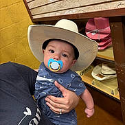 Eastyn is registered to the contest to win money with this photo: accessory, adult_hand, baby, blue_clothing, casual, child, clothing, cowboy_hat, cute, hand, hat, holding, indoor, infant, pacifier, person, portrait, shelf, store, yellow_wall