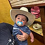 baby, pacifier, cowboy_hat, child, holding, adult_hand, blue_clothing, indoor, shelf, hat, yellow_wall, person, portrait, cute, infant, clothing, hand, accessory, store, casual