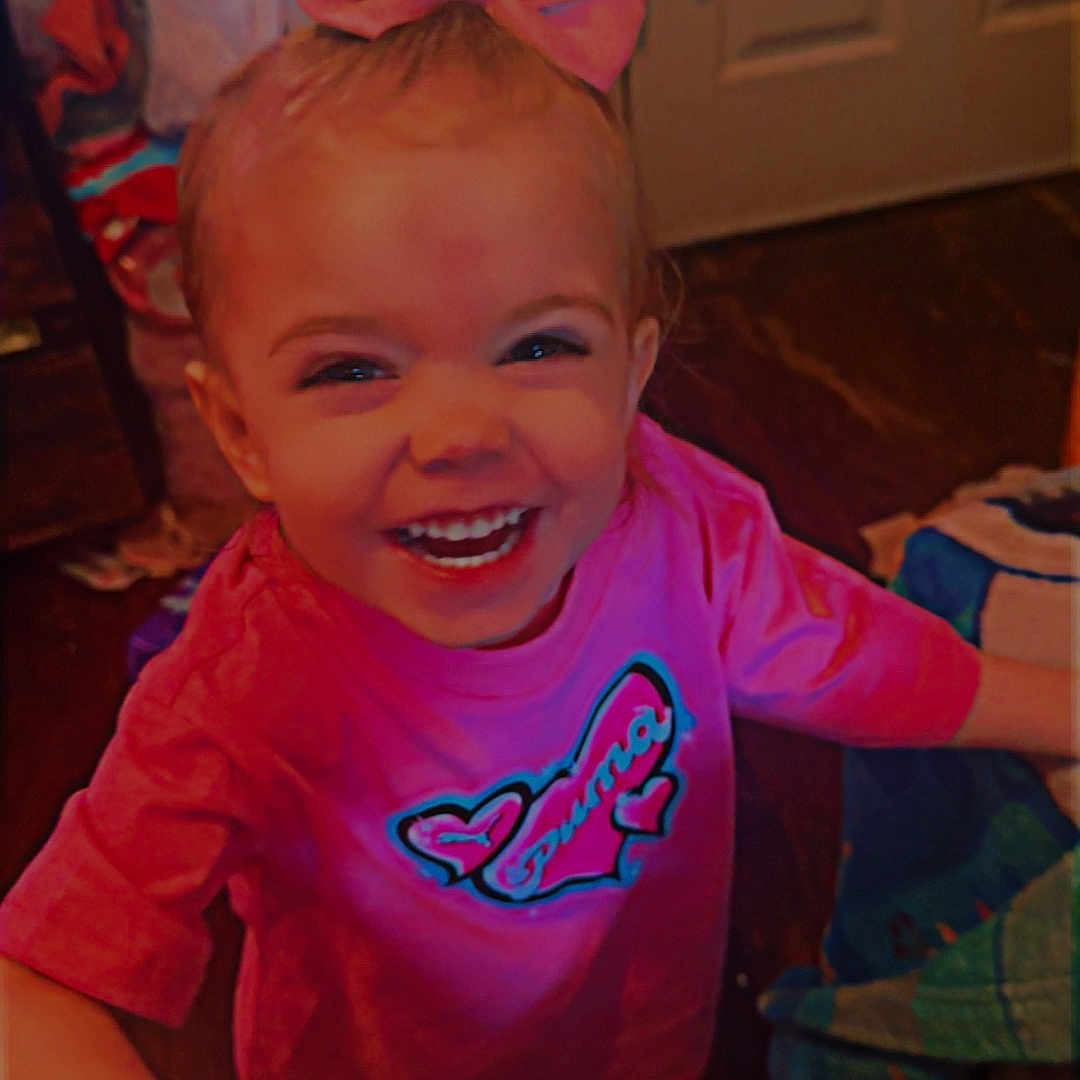 Abagail is registered to the contest to win money with this photo: blanket, child, clothing, door, expression, face, footwear, hair, hand, happy, head, indoor, person, pink_bow, pink_shirt, room, smiling, toddler, toy, wood_floor