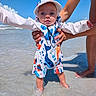 baby, child, beach, ocean, seawater, shoreline, standing, smiling, face, infant, sunny, outdoors, vacation, adult, hands, hat, sunglasses, colorful_clothing, sea_animals, portrait