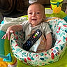 baby, child, infant, smiling, sitting, activity_center, baby_toy, toy_fish, remote_control, colorful, playroom, indoor, portrait, face, hands, gray_shirt, patterned_seat, green, yellow, happy