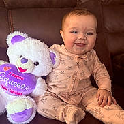 Evelynn is registered to the contest to win money with this photo: baby, infant, smiling, sitting, teddy_bear, stuffed_animal, plush_toy, onesie, pink_clothing, purple, heart_pillow, couch, portrait, face, blue_eyes, hand, soft_texture, indoor, happy, family_moment