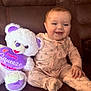 baby, infant, smiling, sitting, teddy_bear, stuffed_animal, plush_toy, onesie, pink_clothing, purple, heart_pillow, couch, portrait, face, blue_eyes, hand, soft_texture, indoor, happy, family_moment