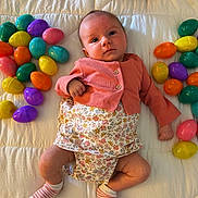 Alaina is registered to the contest to win money with this photo: baby, infant, child, plastic_eggs, easter_eggs, colorful, pink_cardigan, floral_clothing, white_quilt, lying_down, cute, indoors, toy, playful, small, portrait, expression, skin, sock, relaxed
