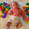 baby, infant, child, plastic_eggs, easter_eggs, colorful, pink_cardigan, floral_clothing, white_quilt, lying_down, cute, indoors, toy, playful, small, portrait, expression, skin, sock, relaxed