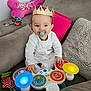 baby, infant, child, pacifier, crown, toy, musical_toy, colorful_lights, sofa, pillow, plush_toy, playtime, hands, eyes, smile, indoors, portrait, happy, seated, home