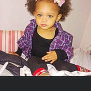 Violet Rose is registered to the contest to win money with this photo: toddler, child, curly_hair, pink_bows, plaid_shirt, black_top, sitting, bed, striped_pillow, indoors, young_child, person, casual_clothing, portrait, cute, looking_at_camera, small_child, light_skin, cozy, home