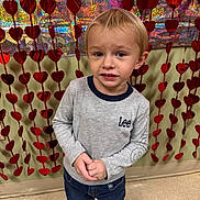 Abel is registered to the contest to win money with this photo: child, toddler, boy, blonde_hair, blue_eyes, smiling, hands_clasped, jeans, long_sleeve_shirt, indoor, heart_garland, red_hearts, sparkly_background, valentine_decor, portrait, standing, tiled_floor, candid_photo, curious_expression, festive