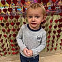 child, toddler, boy, blonde_hair, blue_eyes, smiling, hands_clasped, jeans, long_sleeve_shirt, indoor, heart_garland, red_hearts, sparkly_background, valentine_decor, portrait, standing, tiled_floor, candid_photo, curious_expression, festive