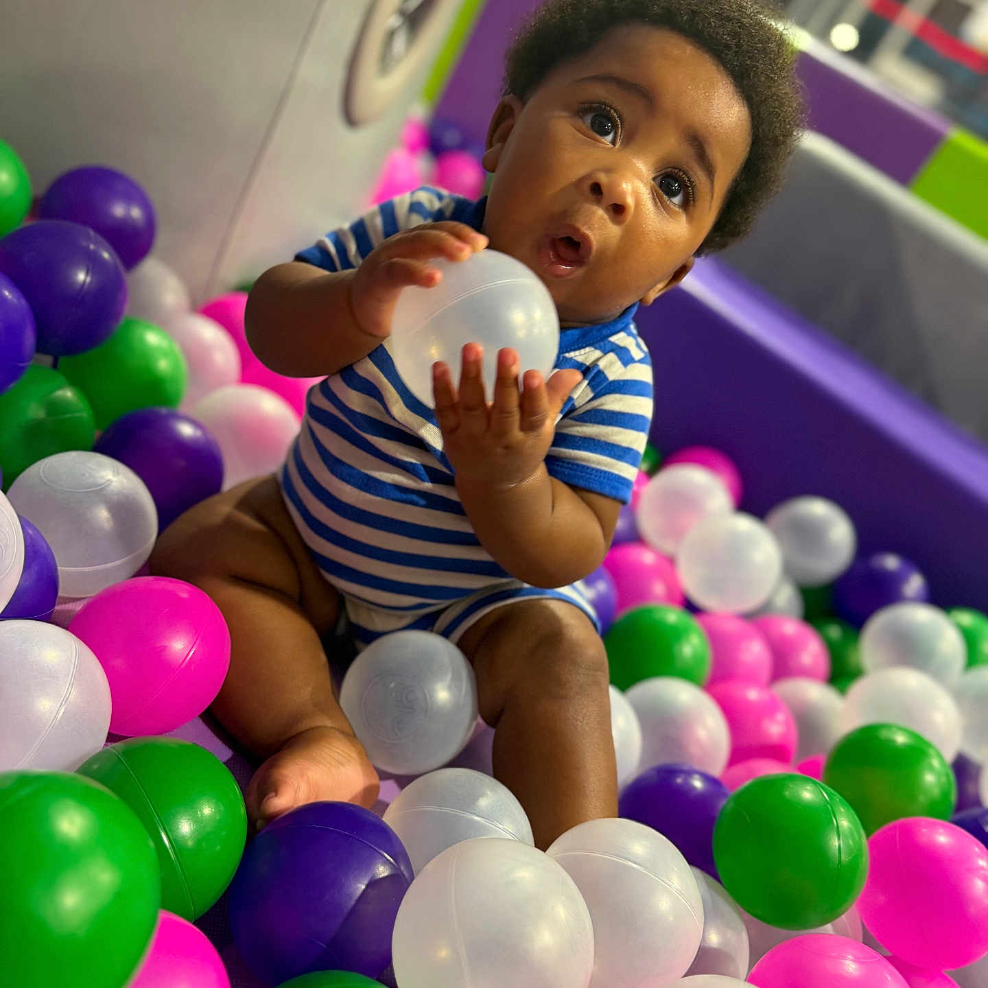 Zymere is registered to the contest to win money with this photo: baby, child, play_area, plastic_balls, striped_onesie, curious, sitting, indoor, colorful, toys, fun, cute, expression, person, head, face, hand, foot, soft_lighting, background_blur