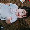 Marcela is registered to the contest to win money with this photo: child, toddler, baby, portrait, smiling, tongue_out, lying_down, carpet, indoor, white_shirt, dark_hair, brown_eyes, fist, playful, toys, cartoon_book, colorful, home, floor, happy