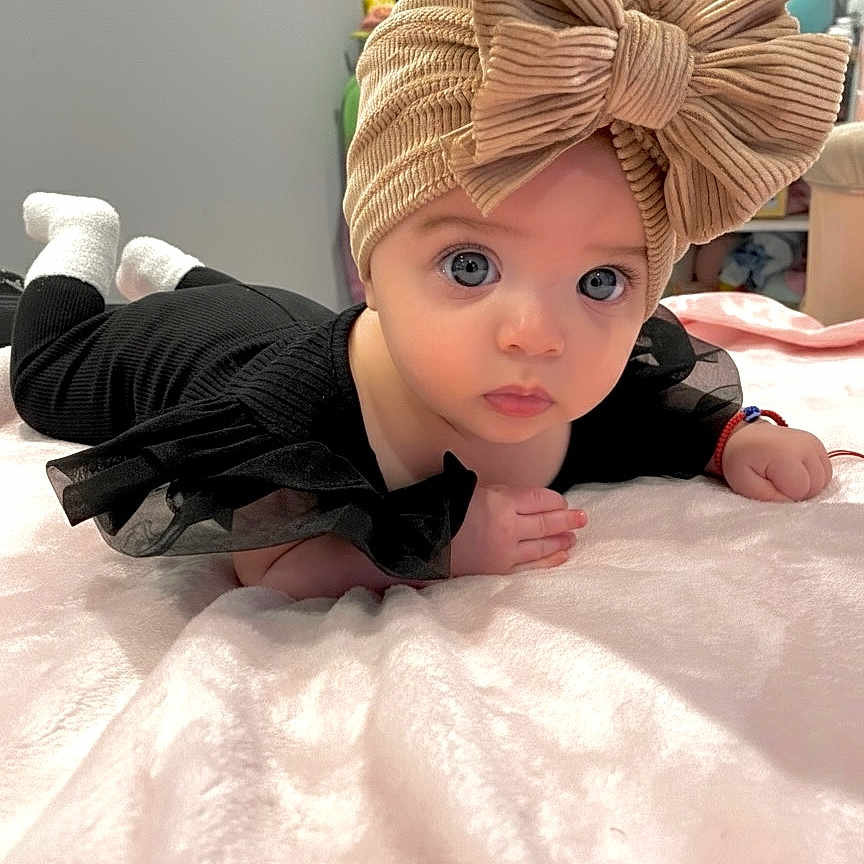 Gabriella joined the competition — help win amazing prizes! baby, black_clothing, blanket, bow, child, cozy, curious, cute, eyes, face, furniture, hand, indoor, infant, lying_down, portrait, room, socks, soft_texture, turban