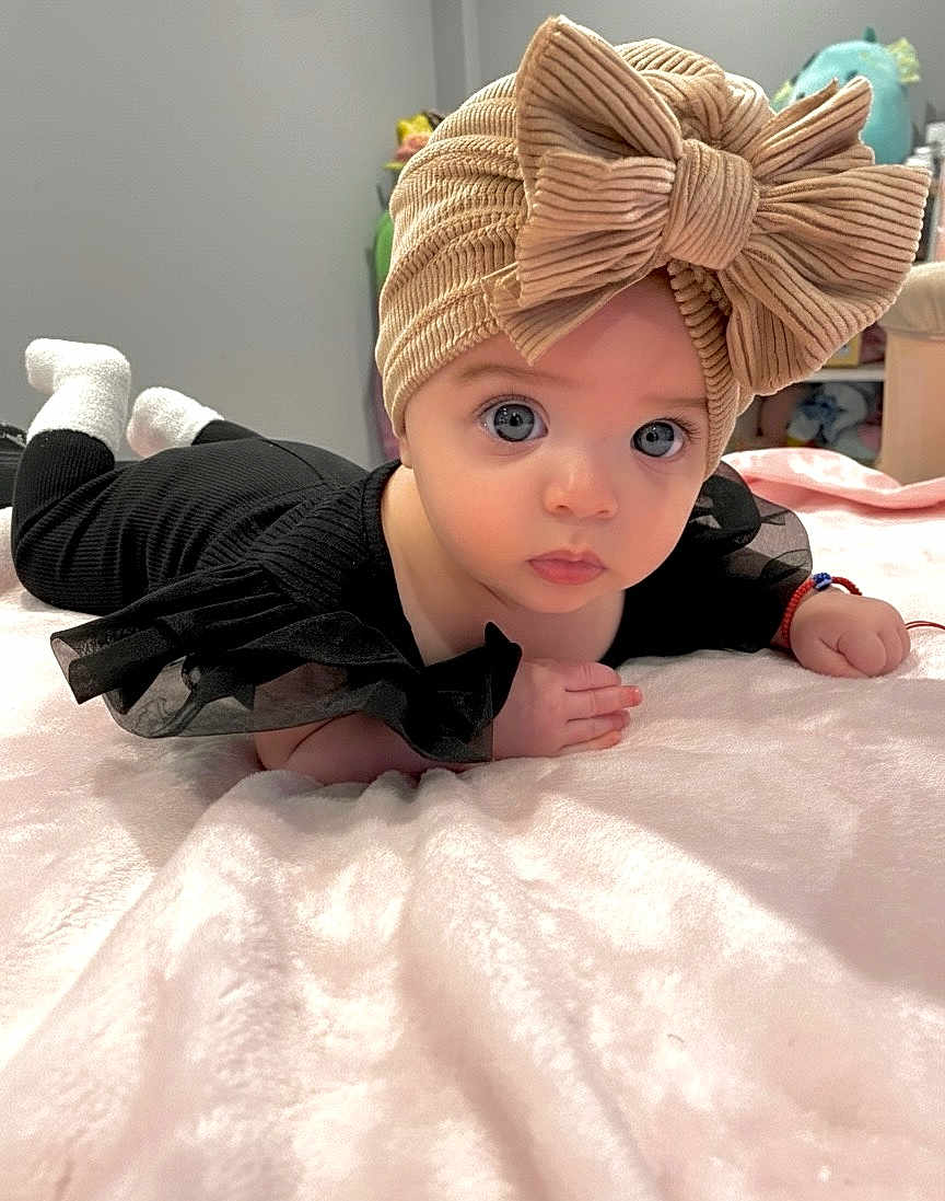 Gabriella joined the competition — help win amazing prizes! baby, child, infant, face, eyes, bow, turban, black_clothing, blanket, soft_texture, indoor, cute, portrait, lying_down, curious, hand, socks, furniture, room, cozy