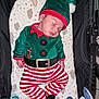 Pierre a rejoint le concours — aidez-le/la à gagner de superbes lots ! baby, sleeping, christmas_elf_costume, hat, striped_pants, baby_cushion, toy, soft_toys, infant, holiday_outfit, cute, child, resting, closed_eyes, indoors, patterned_fabric, black_background, small_hands, footwear, festive