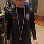 child, boy, medal, sportswear, indoor, smile, short_hair, standing, black_clothing, nike_logo, home, furniture, table, chair, floor, radiator, toy, casual, person, young