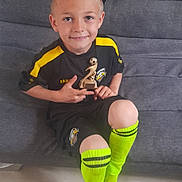 Thyson participe au concours pour gagner de l'argent avec cette photo : child, boy, soccer, trophy, sportswear, yellow_socks, couch, smiling, short_hair, indoor, black_shorts, black_shirt, logo, knees, happy, casual, floor, feet, sitting, portrait