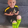 child, boy, soccer, trophy, sportswear, yellow_socks, couch, smiling, short_hair, indoor, black_shorts, black_shirt, logo, knees, happy, casual, floor, feet, sitting, portrait