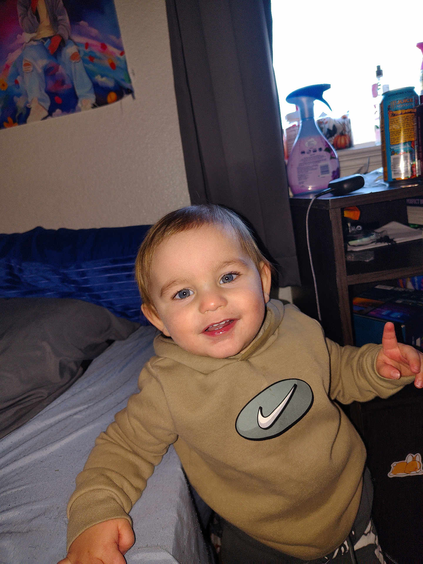 Easton is registered to the contest to win money with this photo: child, toddler, smiling, hoodie, bed, blanket, poster, curtain, nightstand, spray_bottle, can, furniture, indoor, person, face, happy, casual_clothing, light, blue_eyes, hand
