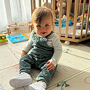 Jules participe au concours pour gagner de l'argent avec cette photo : baby, child, infant, blue_eyes, overalls, white_shoes, play_mat, nursery, crib, toys, indoor, smiling, sitting, cute, soft_light, wooden_furniture, floor, happy, portrait, young_child